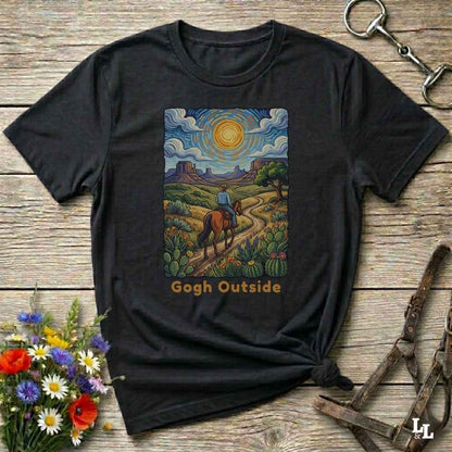 A black graphic t-shirt laid flat on a rustic wooden surface, styled with a small bouquet of flowers, a horse bit, and a leather halter. The t-shirt features a colorful central print depicting a desert landscape with a rider on a horse under a swirling sky, with the text 'Gogh Outside' printed below.