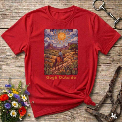 A red short-sleeve graphic T-shirt laid flat on a rustic wooden surface, styled with flowers, a horse bit, and a leather bridle. The shirt has a large rectangular western landscape print on the chest showing a rider on a horse, cacti, mountains, and a sun, with the text 'Gogh Outside' below.