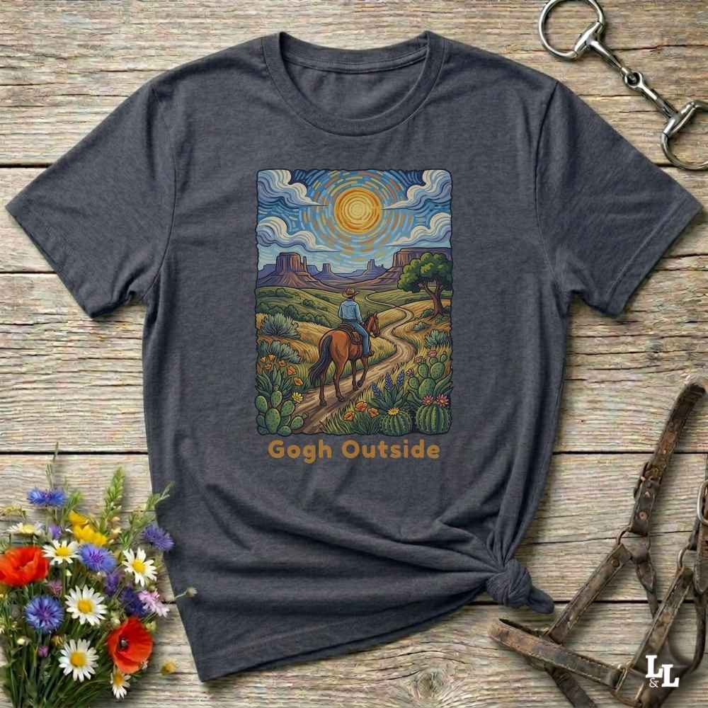 A dark heather gray graphic t-shirt laid flat on a rustic wooden surface, styled with equestrian and floral props. The graphic features a colorful painting of a cowboy riding a horse along a desert path under a swirling sky, with the text 'Gogh Outside' printed below.