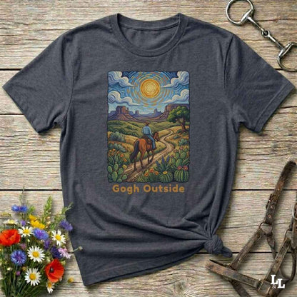 A dark heather gray graphic t-shirt laid flat on a rustic wooden surface, styled with equestrian and floral props. The graphic features a colorful painting of a cowboy riding a horse along a desert path under a swirling sky, with the text 'Gogh Outside' printed below.