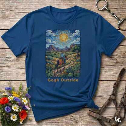 A navy blue graphic t-shirt laid flat on a rustic wooden surface, styled with a horse halter, flowers, and a leather strap. The t-shirt features a colorful art print of a cowboy riding a horse through a desert landscape, with the text 'Gogh Outside' printed below.