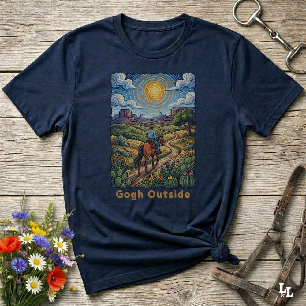 A navy blue graphic t-shirt laid flat on a rustic wooden surface, styled with a small bouquet of flowers, a horse bit, and a leather halter. The t-shirt features a colorful art print of a desert landscape with a rider on a horse and the text 'Gogh Outside'.