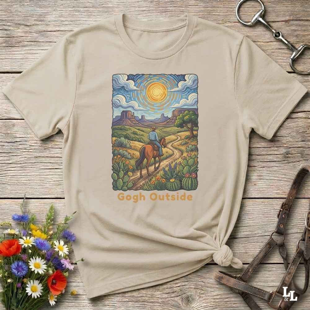 A beige graphic t-shirt laid flat on a rustic wooden surface, styled with a horse bit, leather reins, and a small bouquet of colorful wildflowers. The t-shirt features a central rectangular illustration of a desert landscape with a rider on a horse, cacti, and a bright sun, with the text 'Gogh Outside' printed below.