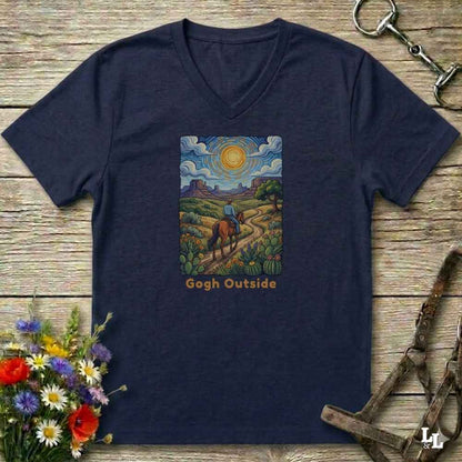 A navy blue V-neck t-shirt laid flat on a rustic wooden surface, decorated with equestrian-themed props like a horse bit, bridle, and a bouquet of wildflowers. The shirt features a colorful graphic print of a rider on a horse in a landscape reminiscent of Gogh's style, with the text 'Gogh Outside' printed below.
