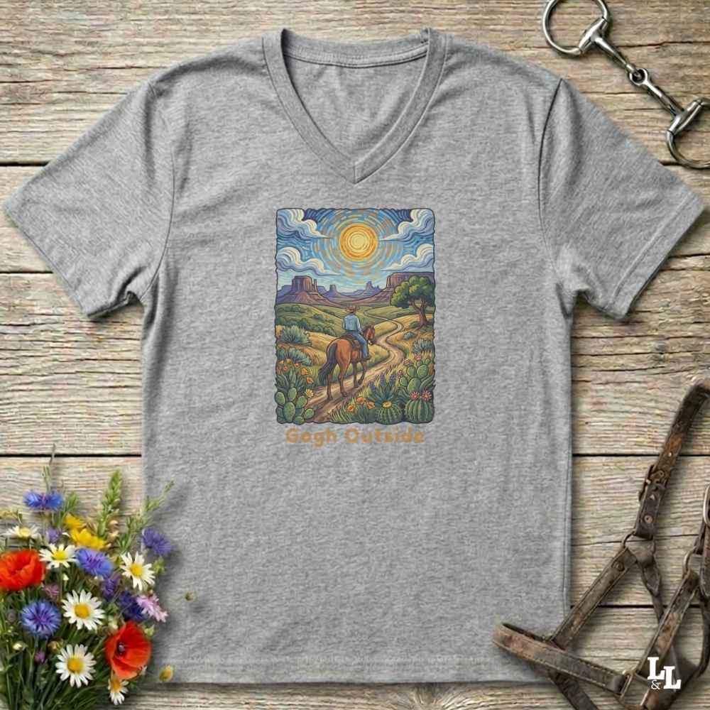 grey V-neck t-shirt laid flat on a rustic wooden surface, featuring a colorful art print of a rider on a horse under a bright sun with the text 'Gogh Outside'. The shirt is styled with equestrian tack and wildflowers.