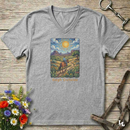 grey V-neck t-shirt laid flat on a rustic wooden surface, featuring a colorful art print of a rider on a horse under a bright sun with the text 'Gogh Outside'. The shirt is styled with equestrian tack and wildflowers.