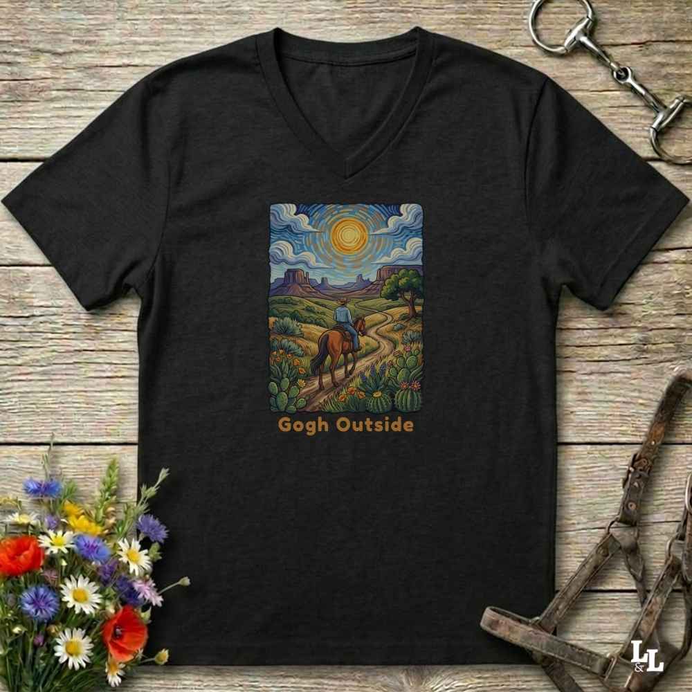 black V-neck t-shirt laid flat on a rustic wooden surface, featuring a colorful art print of a rider on a horse under a bright sun with the text 'Gogh Outside'. The shirt is styled with equestrian tack and wildflowers.