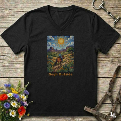 black V-neck t-shirt laid flat on a rustic wooden surface, featuring a colorful art print of a rider on a horse under a bright sun with the text 'Gogh Outside'. The shirt is styled with equestrian tack and wildflowers.