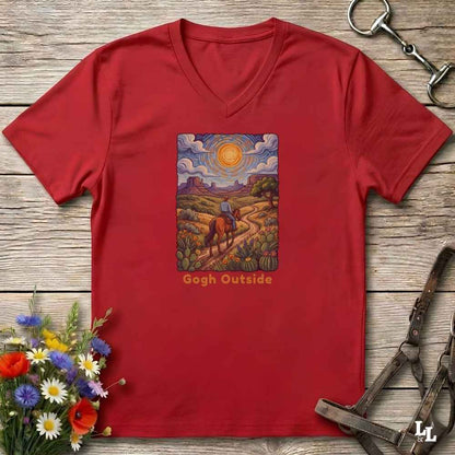 red V-neck t-shirt laid flat on a rustic wooden surface, featuring a colorful art print of a rider on a horse under a bright sun with the text 'Gogh Outside'. The shirt is styled with equestrian tack and wildflowers.