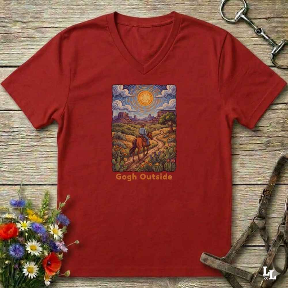 A red heather V-neck t-shirt laid flat on a rustic wooden surface, featuring a colorful art print of a cowboy riding through a landscape in a Van Gogh style, with the text 'Gogh Outside' below the artwork. The shirt is styled with equestrian tack and a flower bouquet.