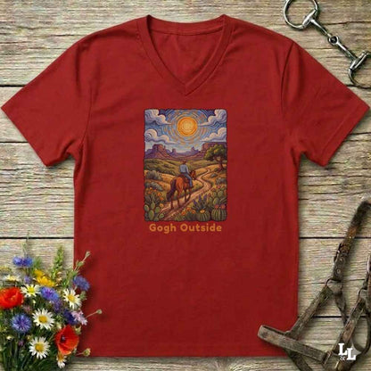 A red heather V-neck t-shirt laid flat on a rustic wooden surface, featuring a colorful art print of a cowboy riding through a landscape in a Van Gogh style, with the text 'Gogh Outside' below the artwork. The shirt is styled with equestrian tack and a flower bouquet.