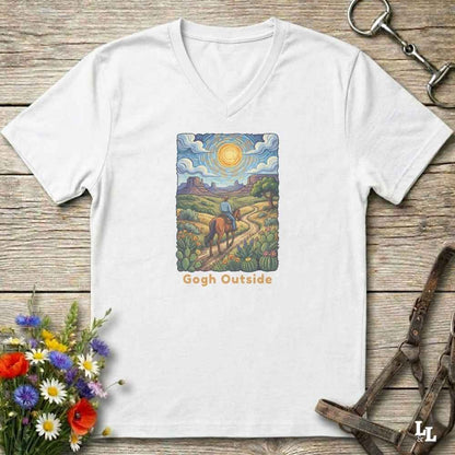 white V-neck t-shirt laid flat on a rustic wooden surface, featuring a colorful art print of a rider on a horse under a bright sun with the text 'Gogh Outside'. The shirt is styled with equestrian tack and wildflowers.