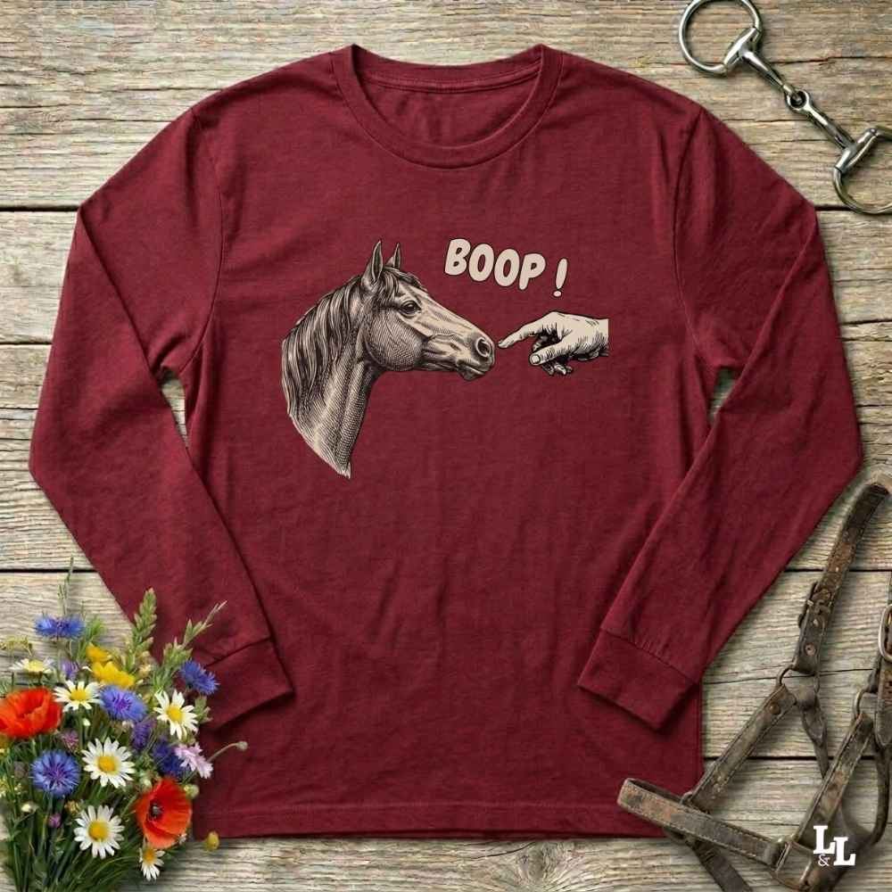 Red long-sleeve shirt with horse graphic and 'BOOP!' text on a wooden surface with flowers and equestrian equipment.