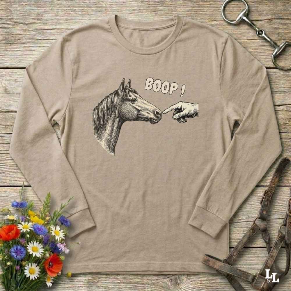 Beige long-sleeve shirt with horse graphic and 'BOOP!' text on a wooden surface with floral and equestrian accessories.