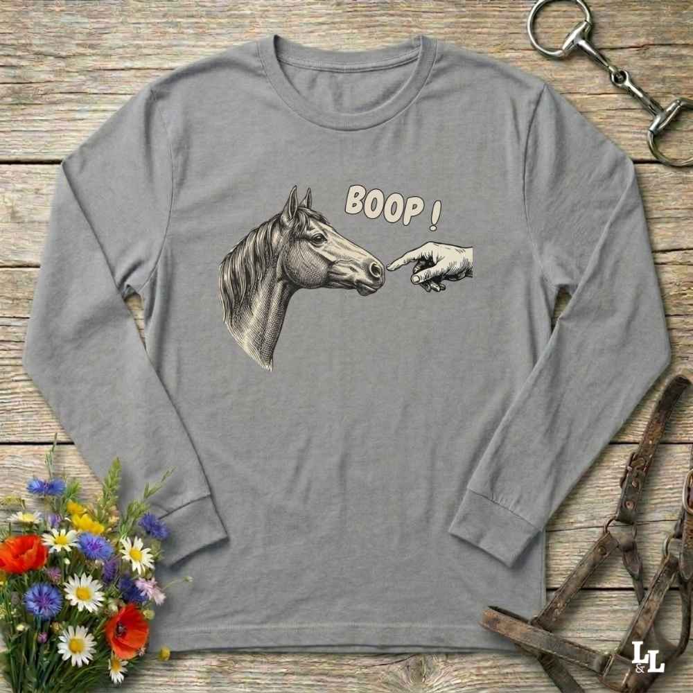 Gray long-sleeve shirt with horse and hand graphic and 'BOOP!' text on a wooden surface with flowers and equestrian equipment.