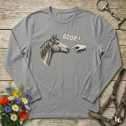 Gray long-sleeve shirt with horse and hand graphic and 'BOOP!' text on a wooden surface with flowers and equestrian equipment.