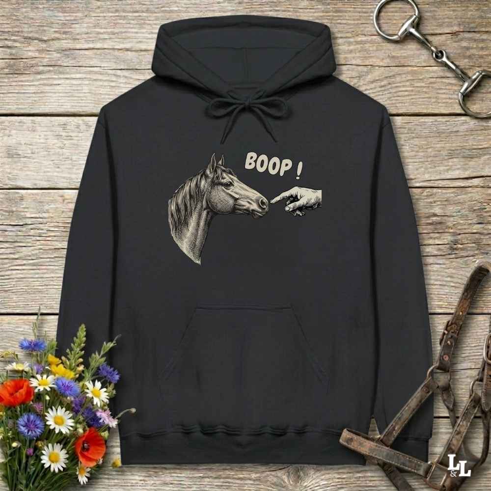 A black pullover hoodie laid flat on a rustic wooden surface, featuring a graphic of a horse head and a pointing hand with the text 'BOOP!' on the chest. The hoodie is styled with equestrian-themed props including a halter, tack, and a small bouquet of flowers.