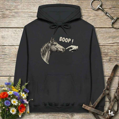 A black pullover hoodie laid flat on a rustic wooden surface, featuring a graphic of a horse head and a pointing hand with the text 'BOOP!' on the chest. The hoodie is styled with equestrian-themed props including a halter, tack, and a small bouquet of flowers.