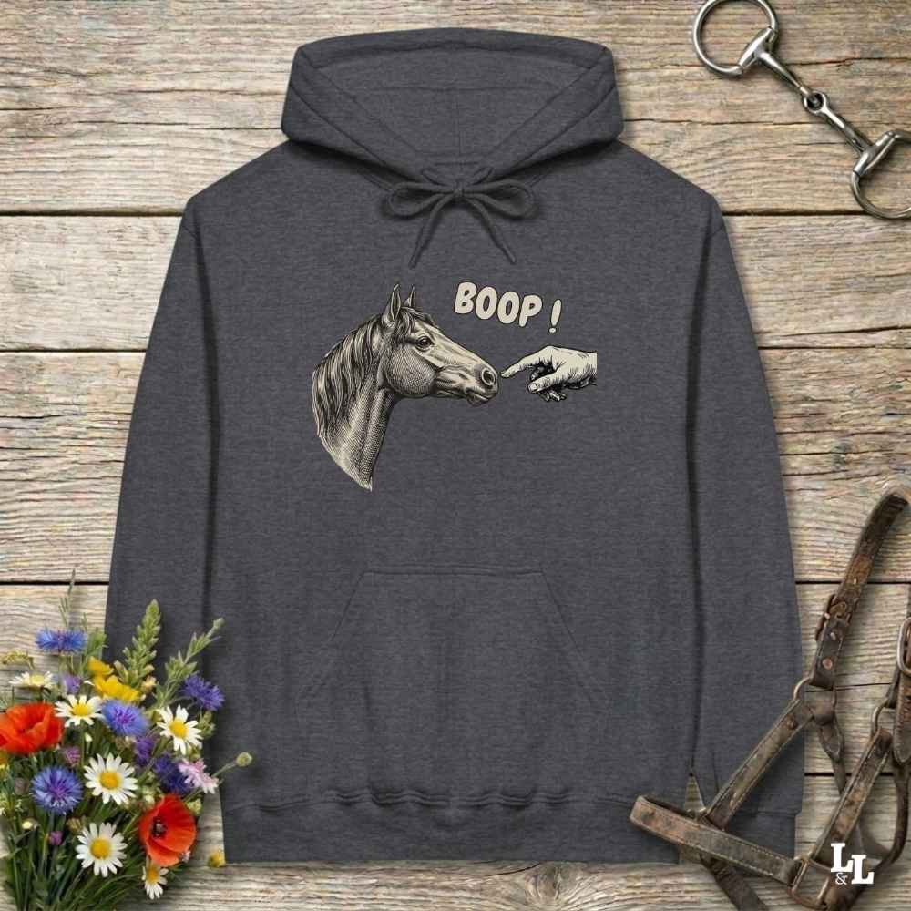 A dark gray hoodie laid flat on a rustic wooden surface, featuring a graphic of a horse head and a pointing hand with the text 'BOOP!'. The hoodie is styled with equestrian-themed props including a halter, tack, and a small bouquet of wildflowers.
