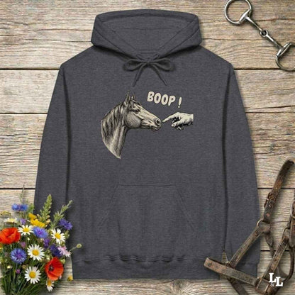 A dark gray hoodie laid flat on a rustic wooden surface, featuring a graphic of a horse head and a pointing hand with the text 'BOOP!'. The hoodie is styled with equestrian-themed props including a halter, tack, and a small bouquet of wildflowers.
