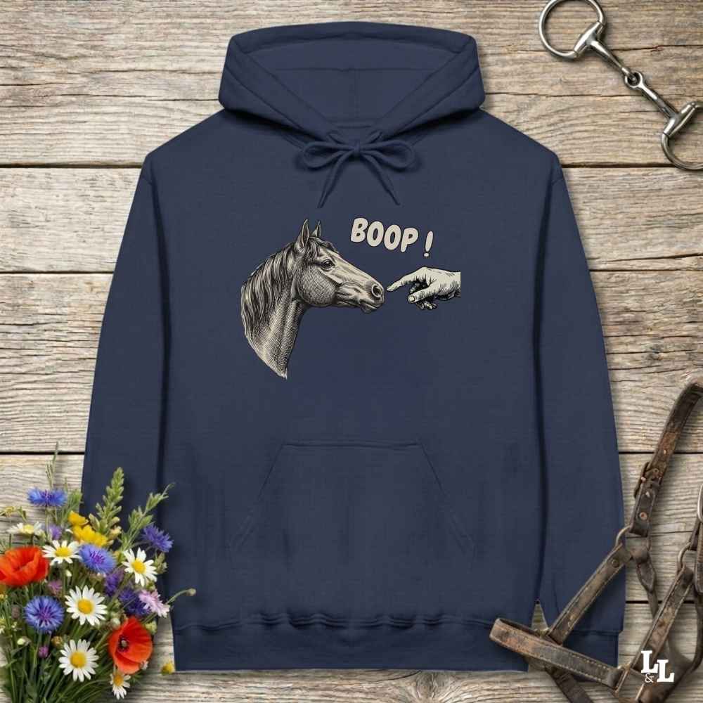 A navy blue hoodie laid flat on a rustic wooden surface, featuring a graphic of a horse head and a pointing hand with the text 'BOOP!' on the chest. The hoodie is styled with equestrian-themed props including a halter, a horse bit, and a small bouquet of flowers.