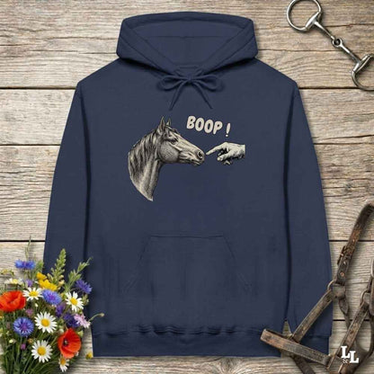 A navy blue hoodie laid flat on a rustic wooden surface, featuring a graphic of a horse head and a pointing hand with the text 'BOOP!' on the chest. The hoodie is styled with equestrian-themed props including a halter, a horse bit, and a small bouquet of flowers.