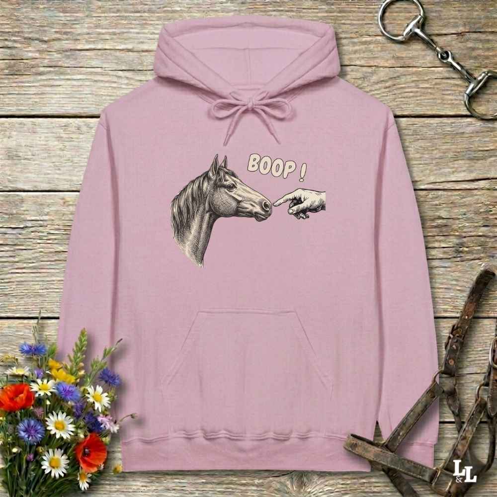 A dusty pink pullover hoodie with a graphic of a horse head and a pointing hand with the text 'BOOP!' on the chest, displayed on a rustic wooden background with equestrian-themed props.