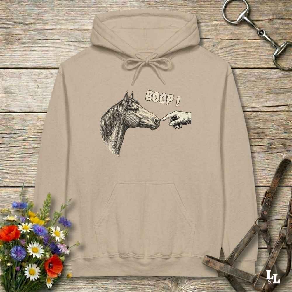 A beige pullover hoodie with a graphic of a horse head and a pointing hand with the text 'BOOP!' on the front, displayed on a rustic wooden background with equestrian-themed props.