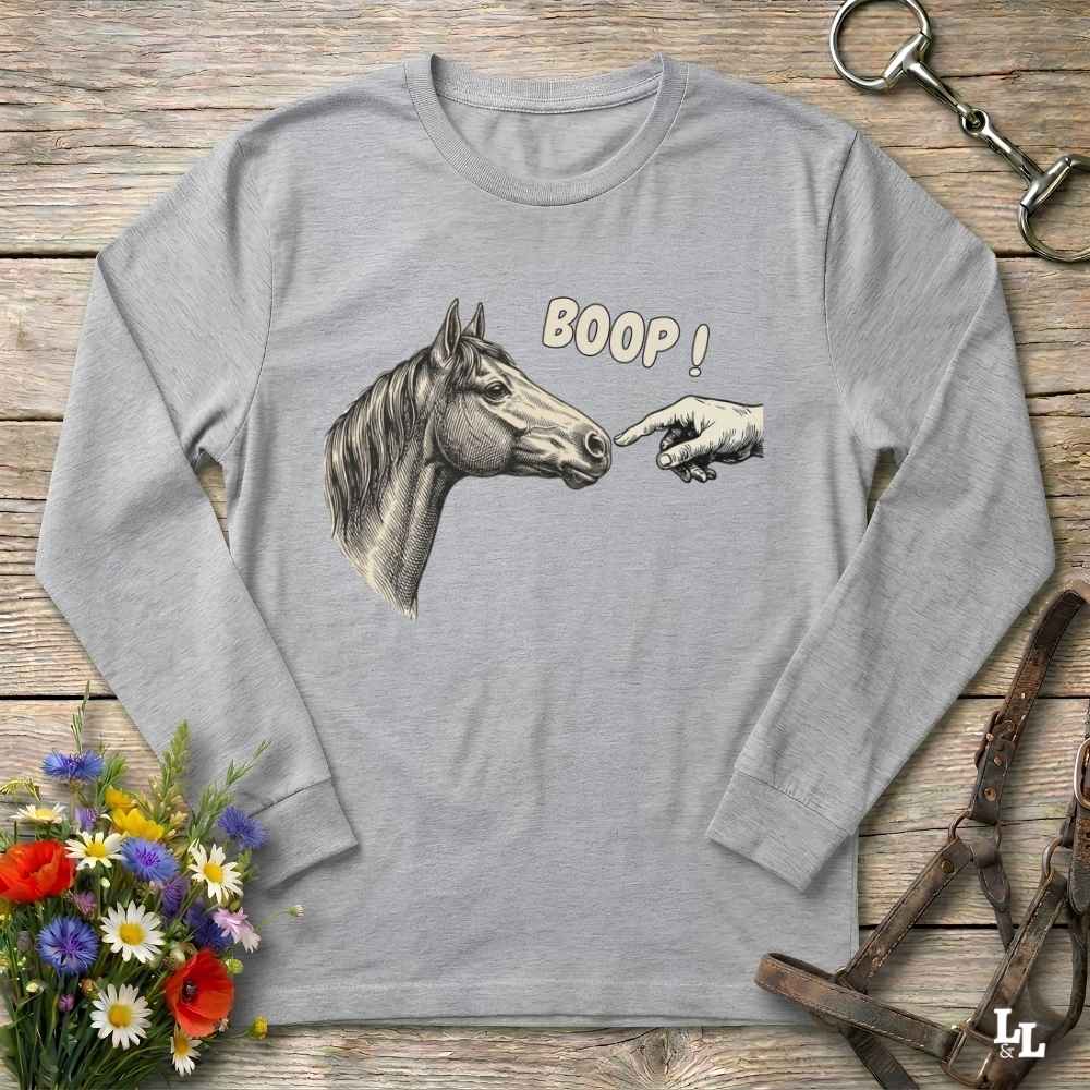 A light gray long-sleeve t-shirt with a graphic of a horse head, a pointing hand, and the text 'BOOP!' printed on the front, displayed flat on a rustic wooden background with decorative equestrian accessories.