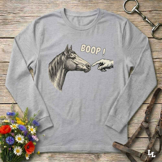 A light gray long-sleeve t-shirt with a graphic of a horse head, a pointing hand, and the text 'BOOP!' printed on the front, displayed flat on a rustic wooden background with decorative equestrian accessories.