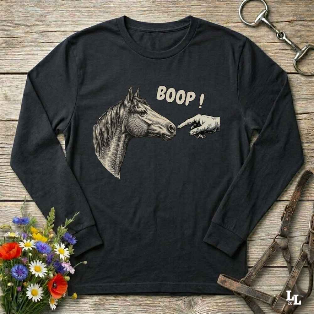 A black long-sleeve t-shirt with a graphic of a horse head and a pointing hand with the text 'BOOP!' printed on the front, displayed on a rustic wooden background with equestrian accessories and flowers.