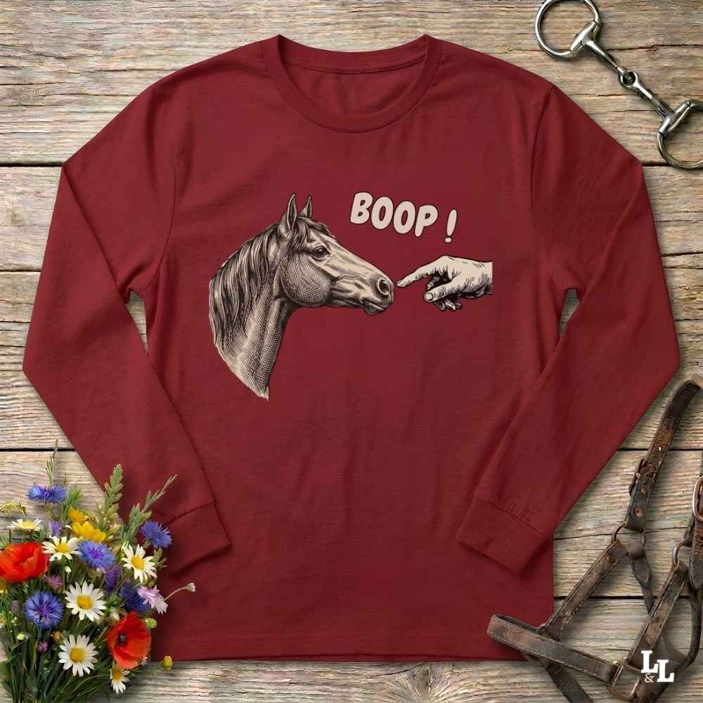 A long-sleeve crewneck shirt in maroon red, featuring a graphic of a horse head facing a pointing hand with the text 'BOOP!'. The shirt is laid flat on a rustic wooden background with equestrian tack and a small bouquet of wildflowers.