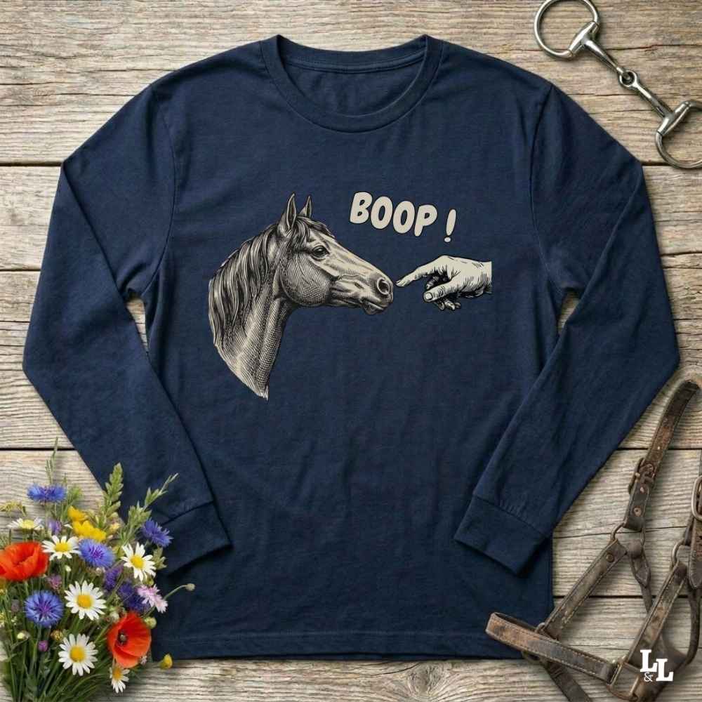 A navy blue long-sleeve t-shirt with a graphic of a horse head and a pointing hand with the text 'BOOP!' printed on the front, displayed on a rustic wooden background with equestrian accessories and flowers.