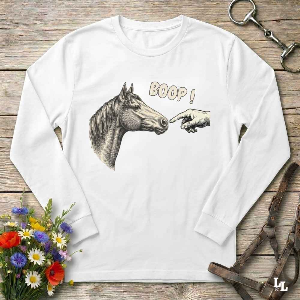 A white long-sleeve t-shirt with a graphic of a horse and a pointing hand with the text 'BOOP!' printed on the front, displayed flat on a rustic wooden background with decorative equestrian and floral props.