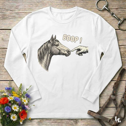A white long-sleeve t-shirt with a graphic of a horse and a pointing hand with the text 'BOOP!' printed on the front, displayed flat on a rustic wooden background with decorative equestrian and floral props.