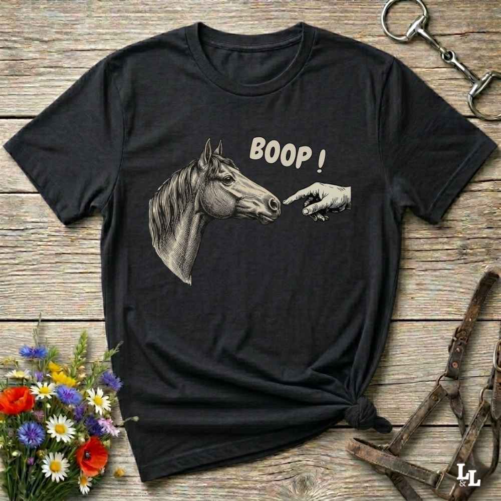 A black short-sleeve graphic t-shirt laid flat on a rustic wooden surface, featuring a grayscale illustration of a horse head pointing at a hand with the text 'BOOP!' in white. The shirt is styled with equestrian and floral props, including a halter, a horse bit, and a small bouquet of wildflowers.