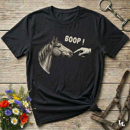 A black short-sleeve graphic t-shirt laid flat on a rustic wooden surface, featuring a grayscale illustration of a horse head pointing at a hand with the text 'BOOP!' in white. The shirt is styled with equestrian and floral props, including a halter, a horse bit, and a small bouquet of wildflowers.