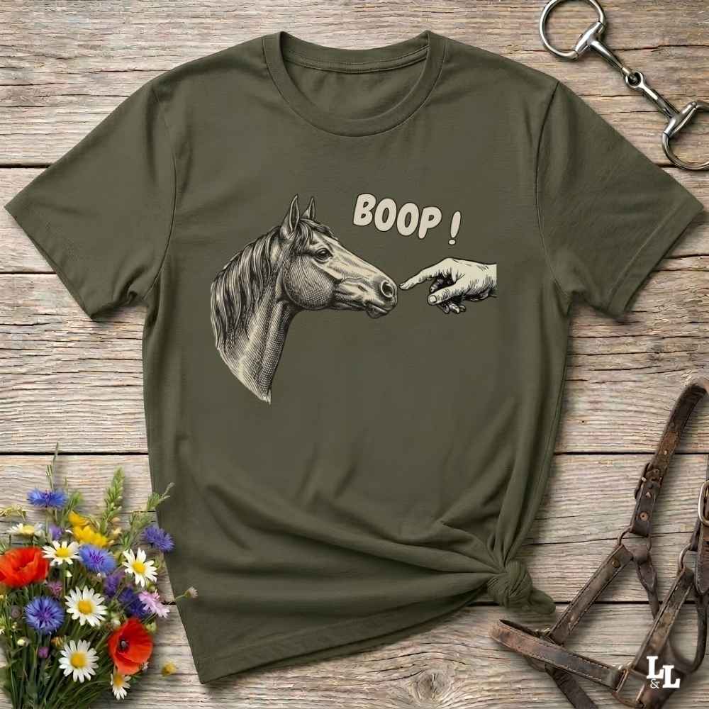 An olive green graphic t-shirt laid flat on a rustic wooden surface, styled with a horse halter, a small bouquet of wildflowers, and a leather bridle. The t-shirt features a graphic of a horse head and a pointing hand with the text 'BOOP!'.
