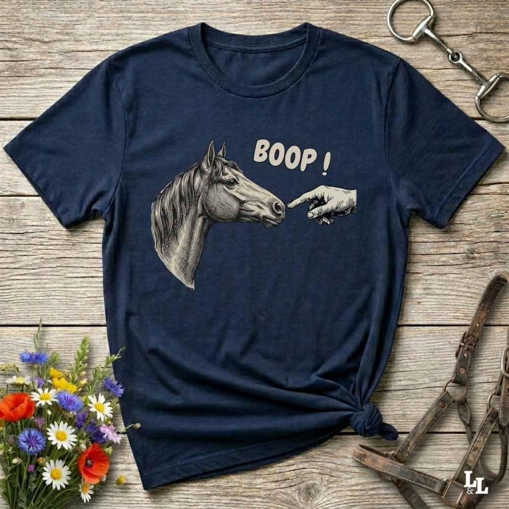 A navy blue graphic t-shirt laid flat on a rustic wooden surface, featuring a grayscale illustration of a horse head and a pointing hand with the text 'BOOP!'. The shirt is styled with equestrian accessories (a halter and a metal bit) and a small bouquet of wildflowers.