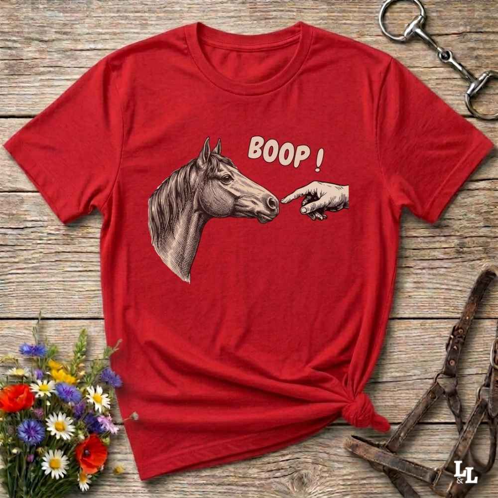 A red short-sleeve graphic T-shirt laid flat on a rustic wooden surface, featuring a horse head, a pointing hand, and the word 'BOOP!' in white. Decorative flowers, a halter, and a metal ring are placed around it as props.