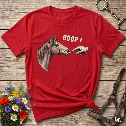 A red short-sleeve graphic T-shirt laid flat on a rustic wooden surface, featuring a horse head, a pointing hand, and the word 'BOOP!' in white. Decorative flowers, a halter, and a metal ring are placed around it as props.