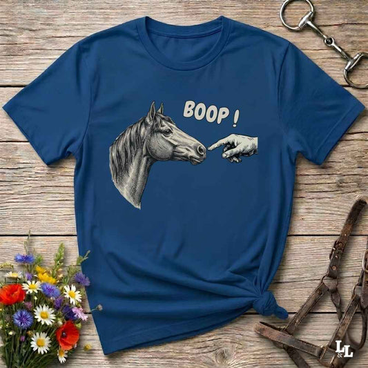 A navy blue graphic t-shirt laid flat on a rustic wooden surface, featuring a grayscale illustration of a horse head and a pointing hand with the text 'BOOP!'. The shirt is styled with equestrian accessories (a leather bridle and a horse bit) and a small bouquet of wildflowers.