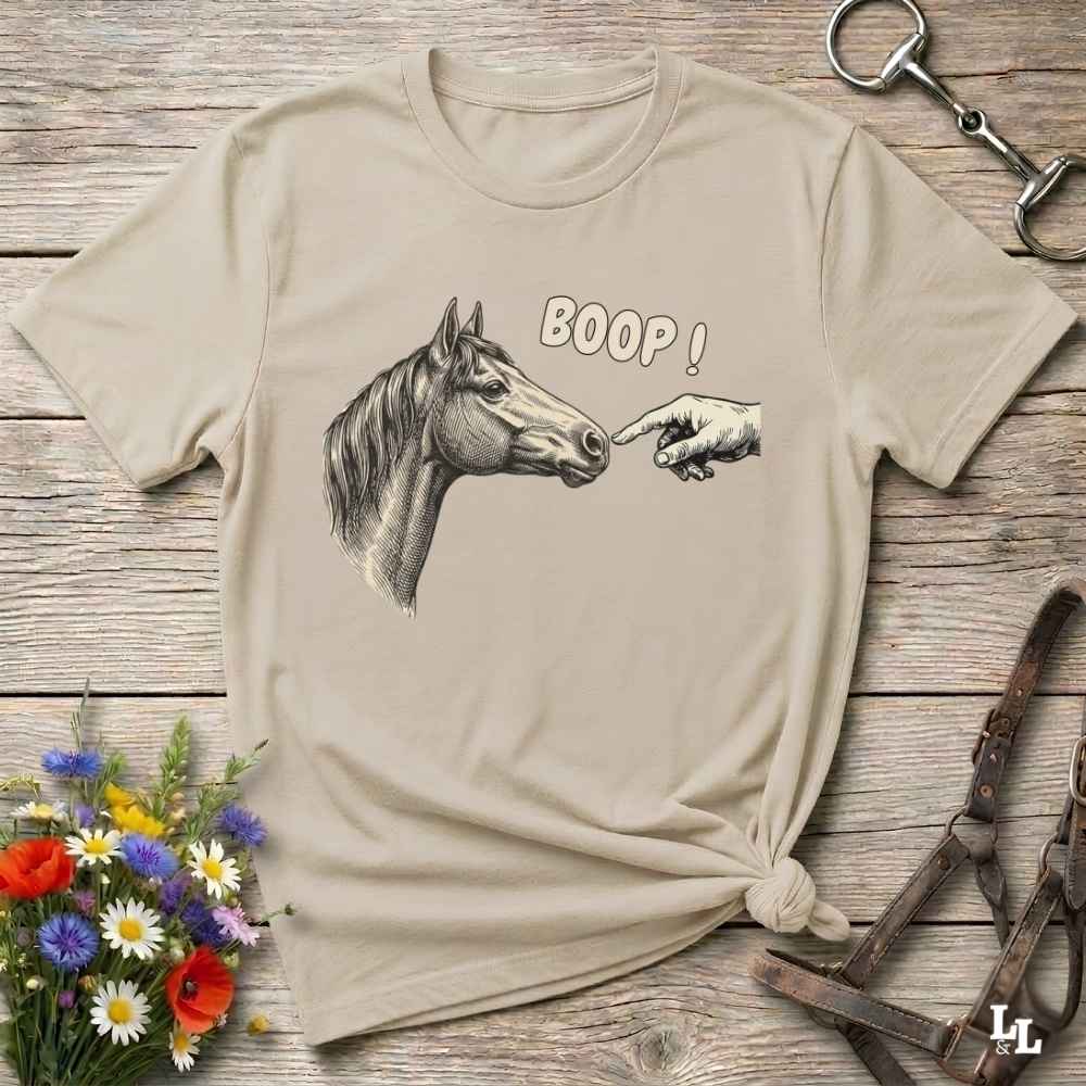 A beige graphic t-shirt featuring a hand pointing at a horse's nose with the text 'BOOP!' printed above it. The t-shirt is styled in a flat-lay arrangement on a rustic wooden background with equestrian accessories and a small bouquet of wildflowers.