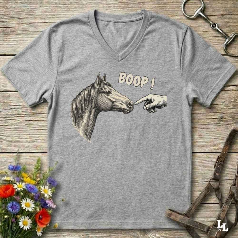 A heather gray V-neck t-shirt laid flat on a rustic wooden surface, featuring a graphic of a horse head and a pointing hand with the text 'BOOP!'. The shirt is styled with equestrian and floral props.