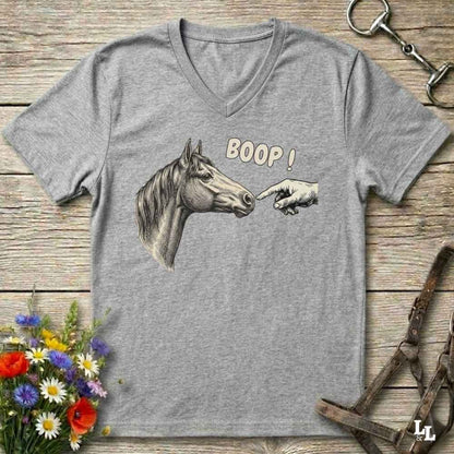 A heather gray V-neck t-shirt laid flat on a rustic wooden surface, featuring a graphic of a horse head and a pointing hand with the text 'BOOP!'. The shirt is styled with equestrian and floral props.