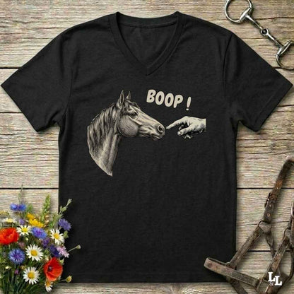 A black V-neck t-shirt with a graphic of a horse head and a pointing hand with the text 'BOOP!' printed on the front, displayed on a rustic wooden background with equestrian accessories and flowers.