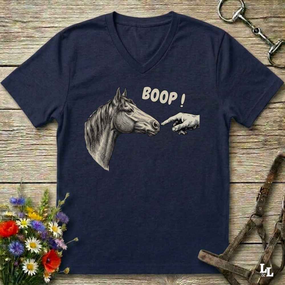 A navy blue V-neck t-shirt laid flat on a rustic wooden surface, featuring a graphic of a horse head and a hand pointing with the text 'BOOP!'. The shirt is styled with equestrian tack, a bouquet of wildflowers, and a metal horse bit.