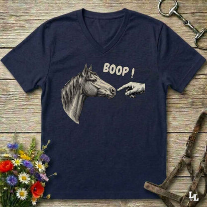 A navy blue V-neck t-shirt laid flat on a rustic wooden surface, featuring a graphic of a horse head and a hand pointing with the text 'BOOP!'. The shirt is styled with equestrian tack, a bouquet of wildflowers, and a metal horse bit.