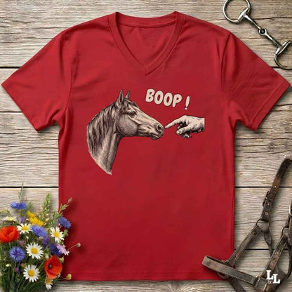 A red V-neck t-shirt laid flat on a rustic wooden surface, featuring a graphic of a horse head and a pointing hand with the text 'BOOP!'. The shirt is styled with equestrian tack and a small bouquet of flowers.