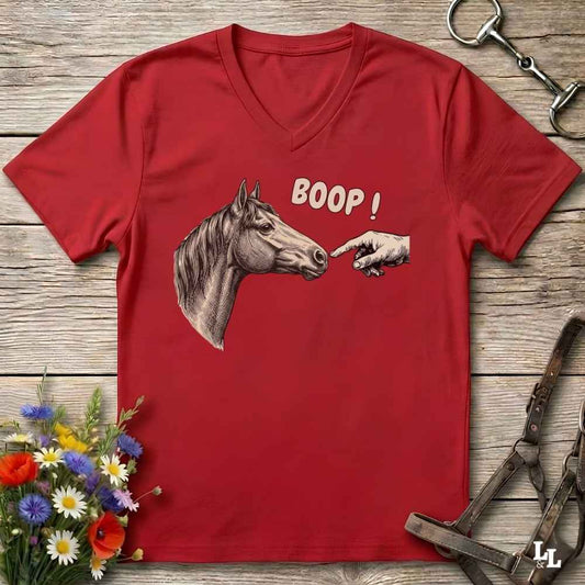 A red V-neck t-shirt laid flat on a rustic wooden surface, featuring a graphic of a horse head and a pointing hand with the text 'BOOP!'. The shirt is styled with equestrian tack and a small bouquet of flowers.